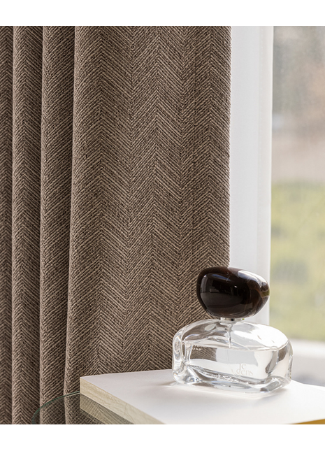 a clear glass perfume bottle with a dark cap, placed on a white surface next to a window with a brown herringbone-patterned curtain.