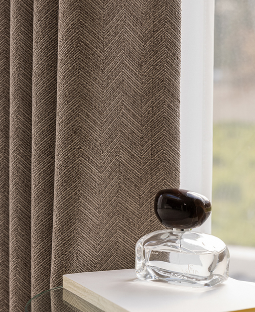 a clear glass perfume bottle with a dark cap, placed on a white surface next to a window with a brown herringbone-patterned curtain.