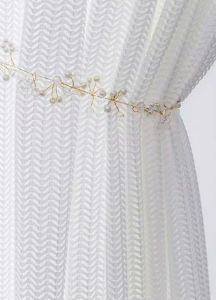 A close-up view of a white, sheer curtain with a decorative gold-colored ribbon or chain attached to it.