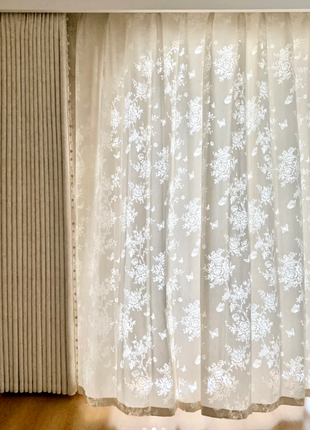 a window with a white curtain featuring a floral pattern. The curtain is partially drawn, allowing some light to enter the room. The curtain appears to be made of sheer fabric, giving it a delicate and elegant appearance.