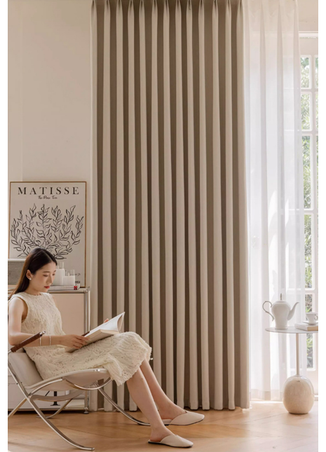 A woman in a white dress is sitting in a chair and reading a book in a room with a large window and a beige curtain.