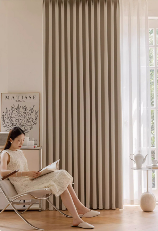 A woman in a white dress is sitting in a chair and reading a book in a room with a large window and a beige curtain.