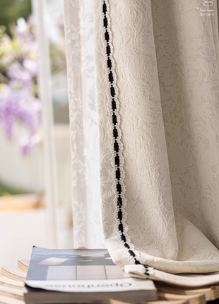 a white curtain with a black lace trim, partially covering a window. On the windowsill, there is a book and a magazine, suggesting a cozy and relaxing atmosphere.
