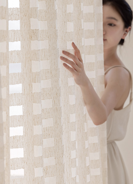 A woman in a white dress stands behind a beige curtain, her hand reaching out to the curtain.