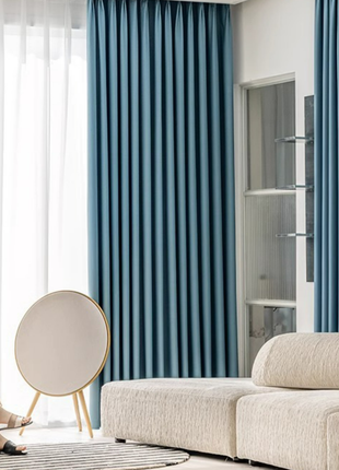 a modern living room with a large window, blue curtains, and a white sofa. A person is sitting on the sofa, and there is a round speaker on a stand in the room.