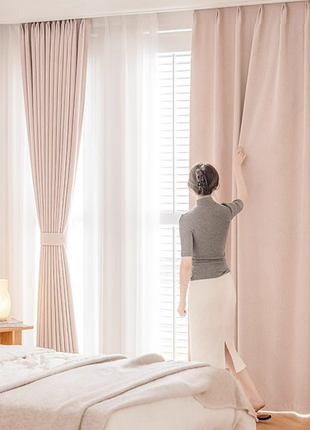 A woman stands in a bedroom, adjusting the curtains on a window.