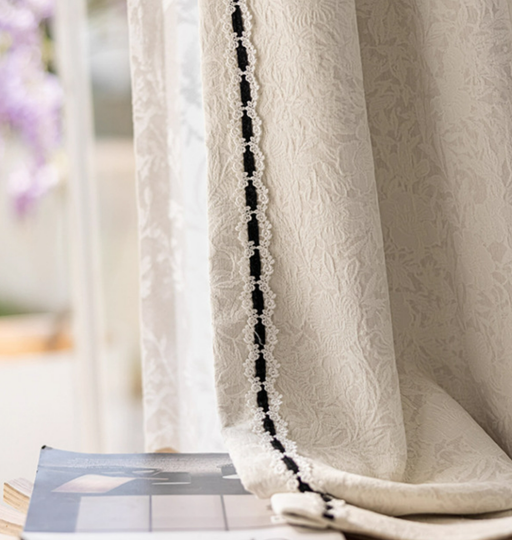 a close-up view of a white curtain with a black lace trim, hanging in front of a window. The curtain appears to be made of a delicate fabric, possibly lace, and is adorned with a decorative black lace trim. The curtain is partially drawn, revealing a glimpse of the window behind it. The background is blurred, drawing attention to the curtain and its details.