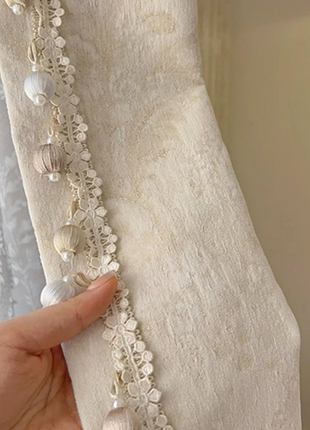 A hand is holding a piece of fabric with a delicate lace trim and small white or beige beads hanging from it.