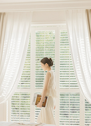 A woman stands in a bedroom, looking out a window with white curtains.