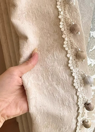 A hand is holding a beige fabric with a decorative trim, featuring small white and brown decorative elements.